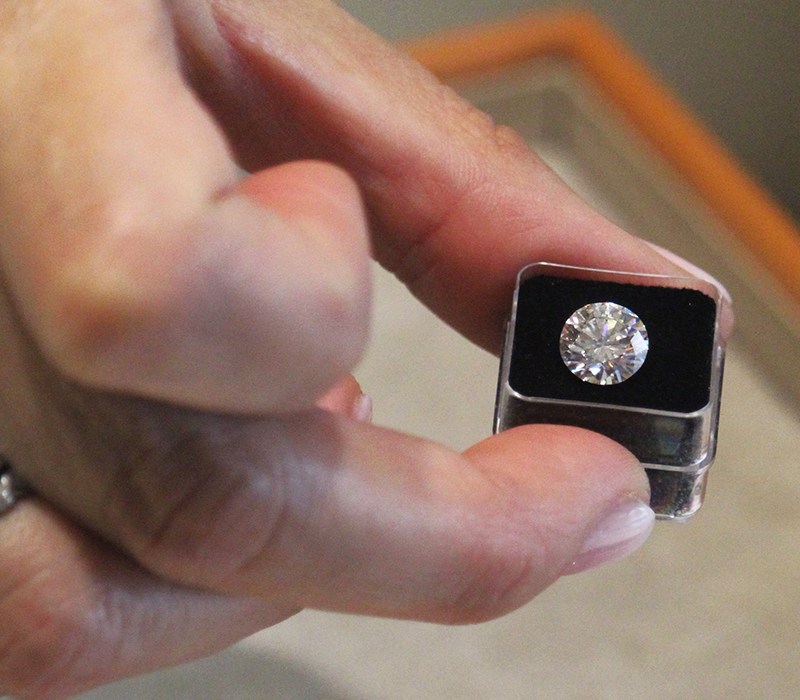 Eine Frauenhand mit auffälligem Diamantenringe übereinander gestapelt hält einen Diamanten in einer durchsichtigen kleinen Box, der auf schwarzem Samtstoff liegt