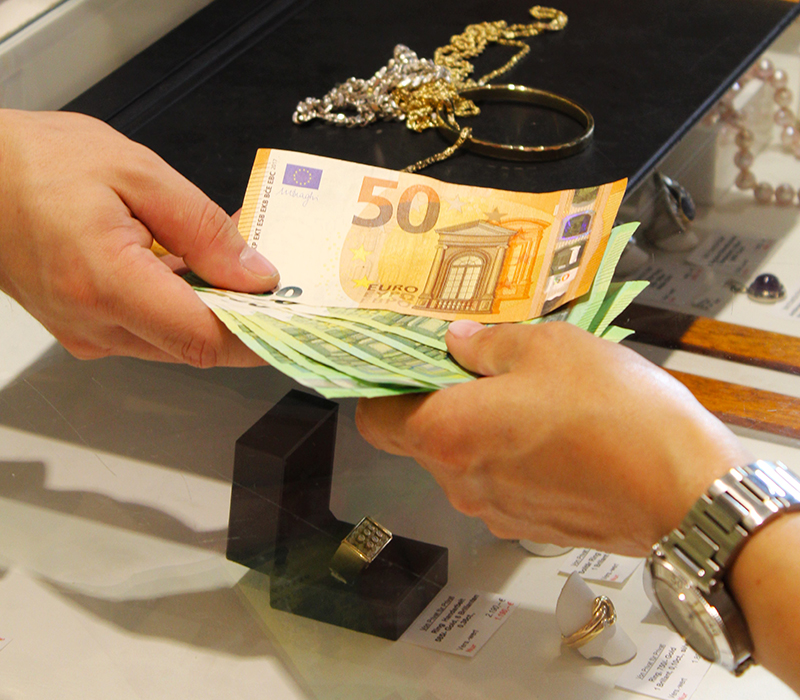 Zwei Hände reichen sich gegenseitig mehrere Geldscheine, im Hintergrund sieht man verschiedenen Schmuckstücke auf einem Präsentiertablett mit schwarzem Samt.