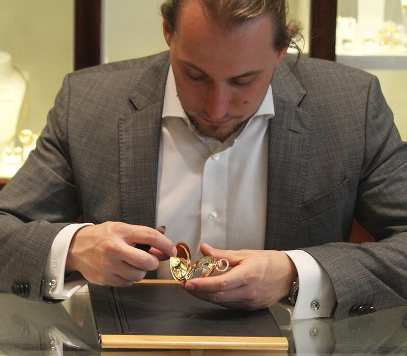 Ein Mann im Anzug sitz an einem Tisch mit Glasplatte und bereitet eine goldenen Taschenuhr auf